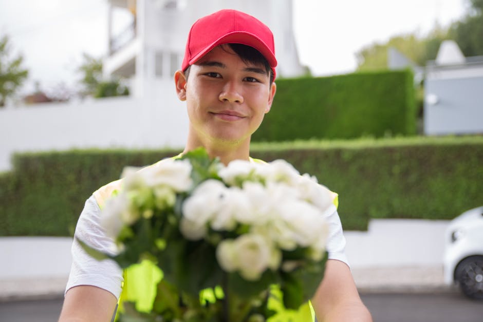 A delivery person wearing a red cap and a neon yellow reflective vest holds a bouquet of fresh white flowers with green leaves, extending it towards the camera in an outdoor residential area. The bouq
