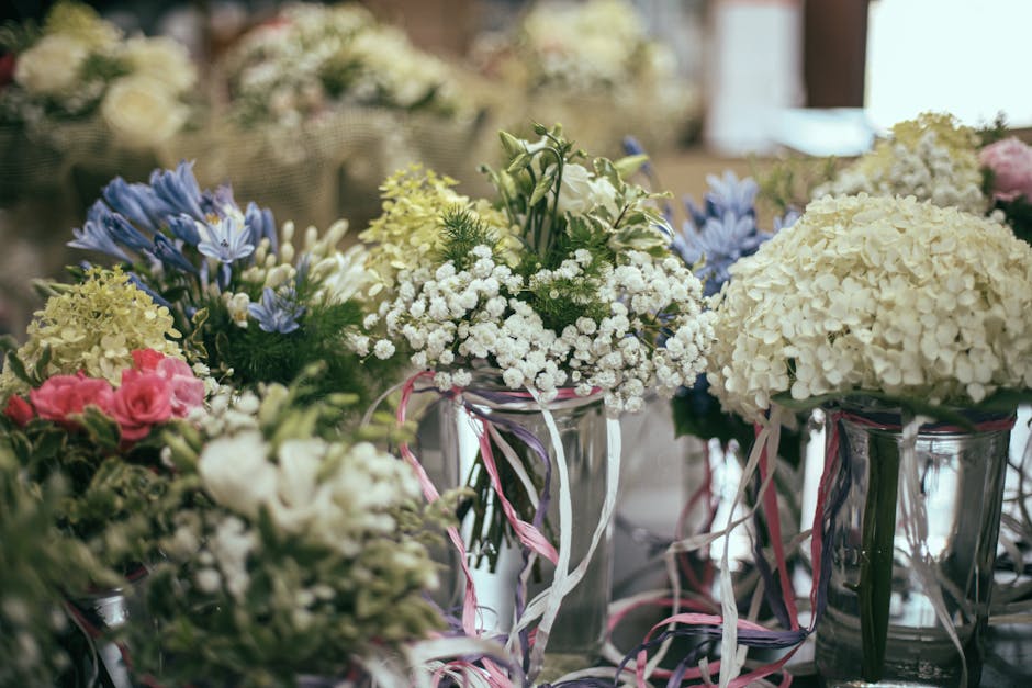 A bouquet of fresh flowers including white hydrangeas, white baby's breath, and pale yellow and pink roses, arranged in clear glass vases decorated with pink and white ribbons. The flowers are placed