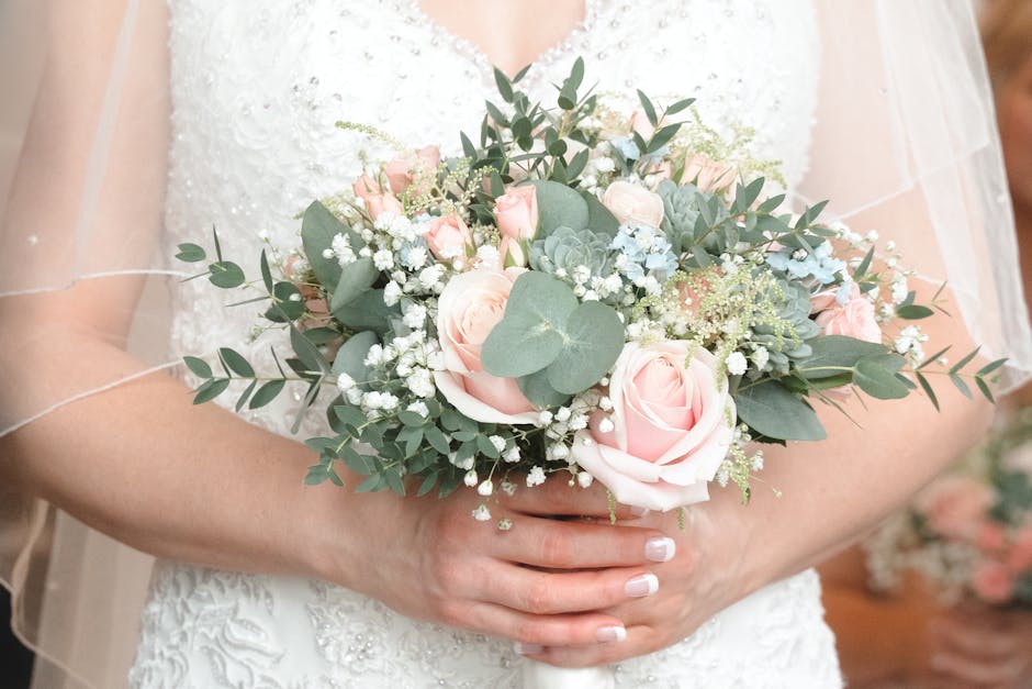 A bride holding a floral bouquet arrangement consisting of soft pink roses, white baby's breath, silvery green eucalyptus, and succulent leaves, in front of her white lace wedding dress. The bouquet i