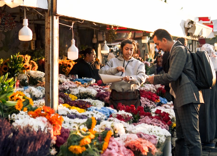 A flower merchant at an outdoor market stand surrounded by an abundance of vibrant, freshly cut flowers including sunflowers, chrysanthemums, roses, and carnations in various bright colours such as ye