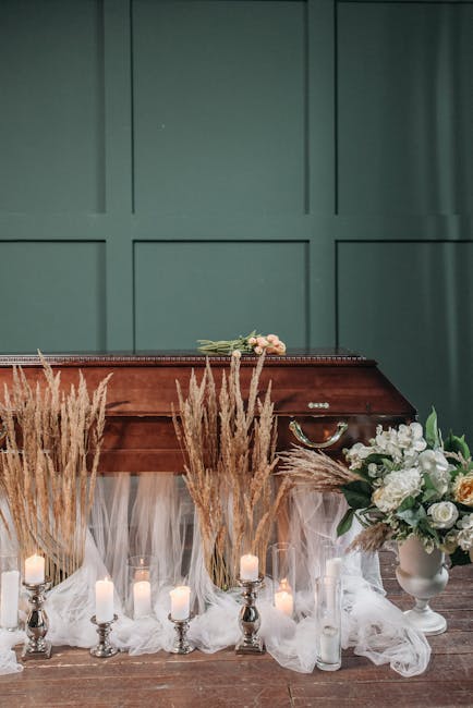 A floral display featuring a bouquet of pale pink roses and white flowers resting on top of a closed wooden casket, surrounded by tall, beige pampas grass and lit candle arrangements placed on a sheer