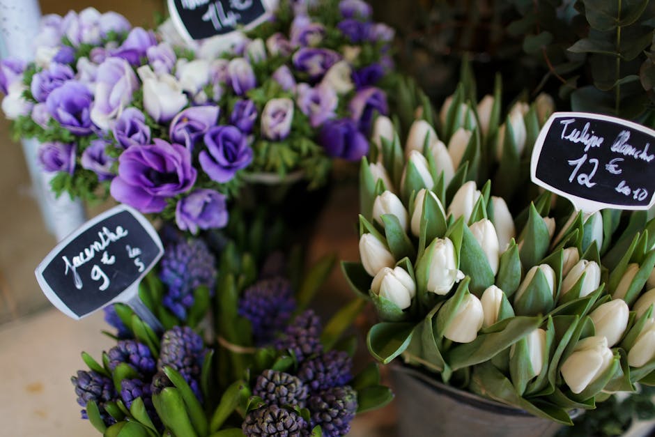 Close-up of a floral display featuring a bouquet of purple and white eustoma (lisianthus) flowers with delicate petals and green leaves, arranged in a black vase. Surrounding the bouquet are clusters