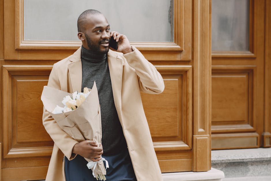 A man with a dark complexion, wearing a beige trench coat over a black turtleneck sweater, stands outside a building with wooden paneling and large windows. He is smiling slightly while talking on a m