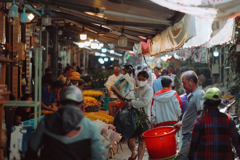 A bustling outdoor flower market at Brixton Market featuring an array of fresh flowers including yellow, orange, and purple blooms, arranged in bouquets on vendor stalls. The scene captures a diverse