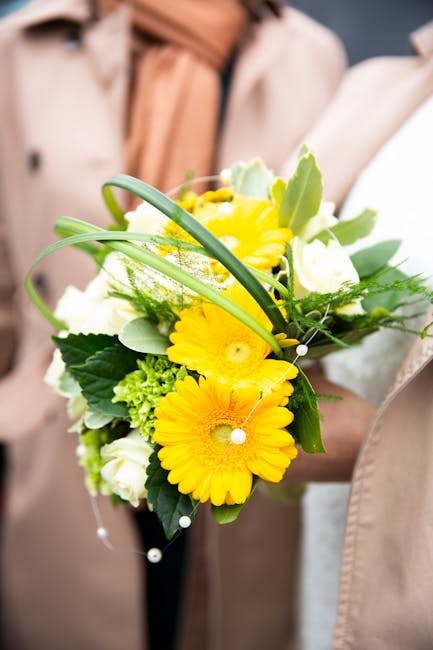 A floral arrangement featuring bright yellow gerbera daisies, complemented by white roses and green foliage, including large leaves and delicate filler greens, all arranged in a compact bouquet. The f
