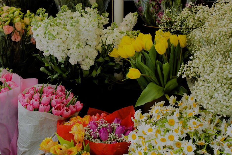 A vibrant display of fresh floral arrangements featuring yellow tulips with long green leaves, white daisies with yellow centers, pink tulips wrapped in pastel pink paper, and clusters of white hydran