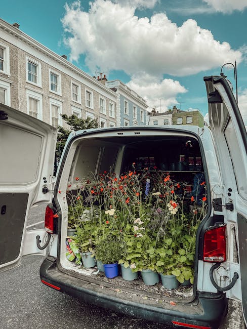 The open rear of a white van reveals an array of potted flowers, predominantly red and white poppies with green foliage, arranged in light blue containers. The flowers are freshly picked, with delicat