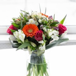Exuding sophistication and warmth, this bouquet is an exquisite reflection of Lambeth's creativity and the artistry of expert local florists. Central to the arrangement, brilliant orange gerbera daisies radiate outward, reminiscent of the vibrant mosaic of cultures that colour Brixton and Oval. Their cheerful faces are softened by the gentle embrace of peach gerberas, whose creamy tones bring a sense of calm and harmony. Deep magenta roses are scattered with intent, their sumptuous spirals offering a depth of feeling akin to cherished memories along the Thames embankment. Toward the heart of the bouquet, delicate sprays of white alstroemeria and pristine spray roses are nestled, their fine textures and crisp colouring illuminating the composition like rays of spring sunlight drifting through Lambeth's historic streets. A wide band of glossy aspidistra leaves curves nobly at the base, echoing the protective canopies found in local gardens. Slender, fresh leaves and textured sprigs fill the spaces between, building density and movement. Presented in a tall, slender glass vase, each verdant stem is visible and full of vitality. Bathed in soft, ambient light, every detail-colour, curve, and petal-gleams. This arrangement evokes celebration and gratitude, whether for an anniversary or simply to remind someone in Lambeth of your appreciation and care.